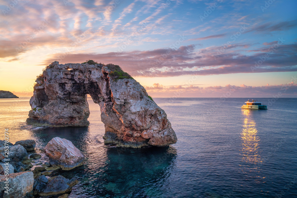 Es Pontas in Mallorca with boat anchored nearby in the Mediterranean sea. Sunrise with sun glinting off the yacht and rock arch.