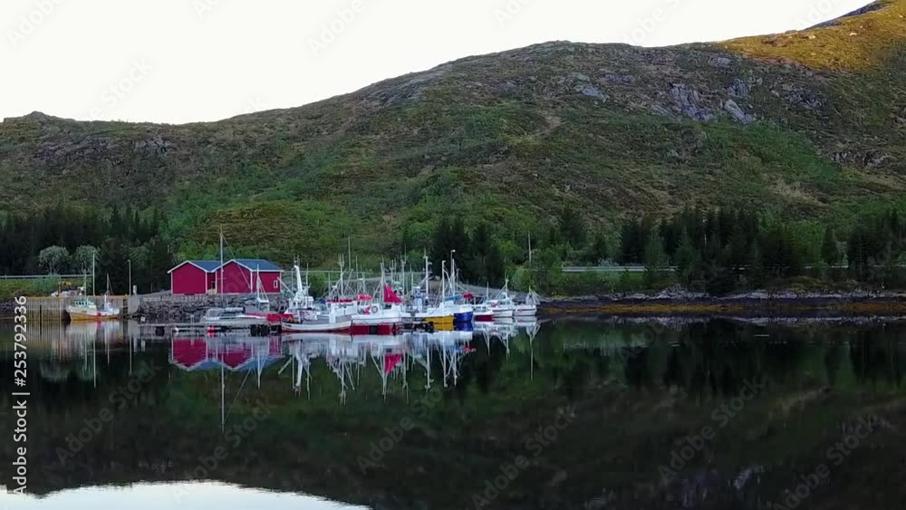 Aerial, drone shot, towards a pier, full of fishing boats, on the coast of Reine town, in the Lofoten islands, in front of mountains, on a sunny, summer evening, in Norway Aerial, drone shot, towards a pier, full of fishing boats, on the coast of Reine town, in the Lofoten islands, in front of mountains, on a sunny, summer evening, in Norway