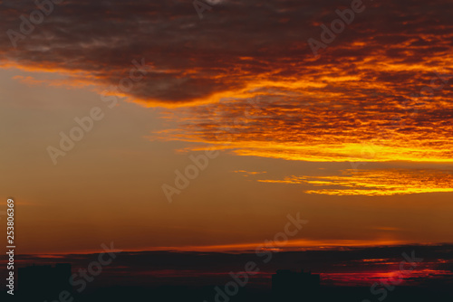 Cityscape with vivid fiery dawn. Amazing warm dramatic cloudy sky above dark silhouettes of city building roofs. Orange sunlight. Atmospheric background of sunrise in overcast weather. Copy space.