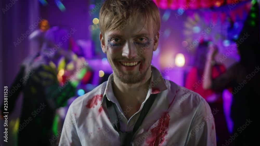 Halloween Costume Party: Portrait of Brain Dead Zombie Wearing Bloody Suit Smiles Creepily. In the Background Monsters Having Fun and Dancing in the Decorated Room Halloween Costume Party: Portrait of Brain Dead Zombie Wearing Bloody Suit Smiles Creepily. In the Background Monsters Having Fun and Dancing in the Decorated Room