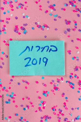 Hebrew text :"Elections 2019" on voting paper over pink with colorful confetti background. Israeli legislative Elections for the 21st Knesset Israel 9 April 2019.