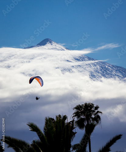 Paraglider flying high among the clouds with a big mountain in the background and palm trees in the foreground