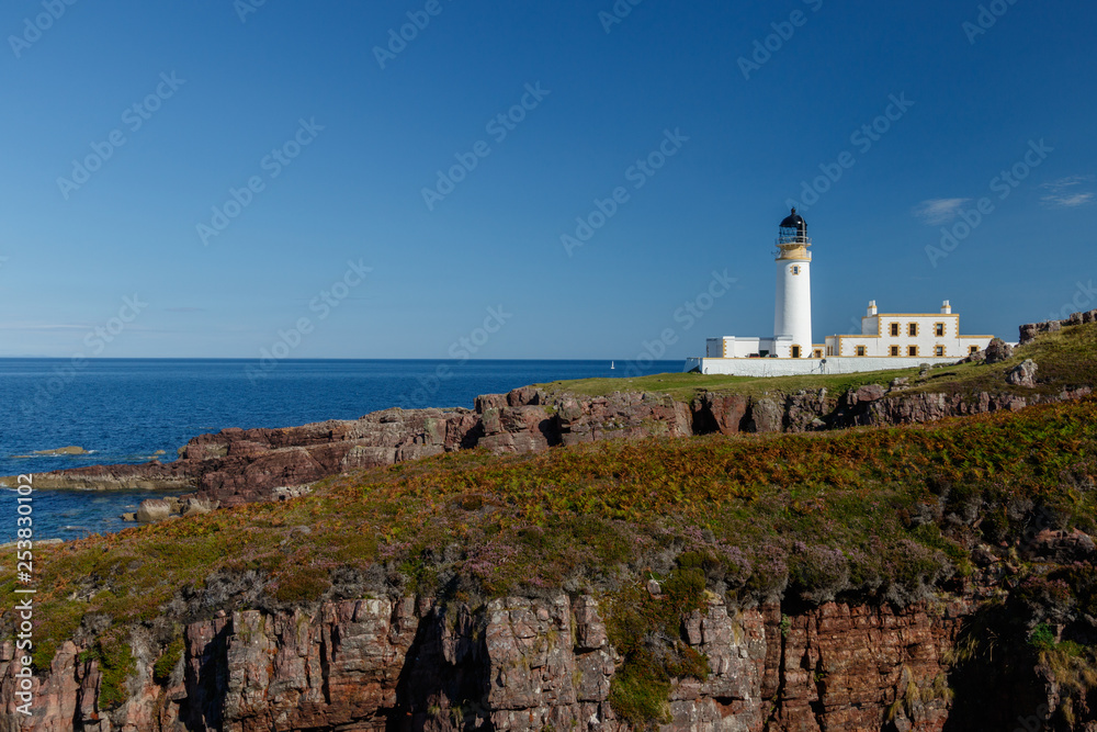 Rua Reidh lighthouse with keepers house on a rocky, grassy coastline with blue sky.