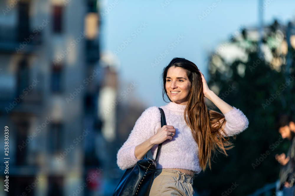 Fototapeta premium Front view of beautiful trendy young woman wearing casual wear sitting on a metallic fence in the street while looking away in a sunny day