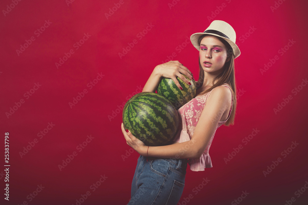 Confident Attractive girl with pink make up, wearing jeans, hat and top, posing at red studio background, holding few watermelon, emotionally stand Confident Attractive girl with pink make up, wearing jeans, hat and top, posing at red studio background, holding few watermelon, emotionally stand