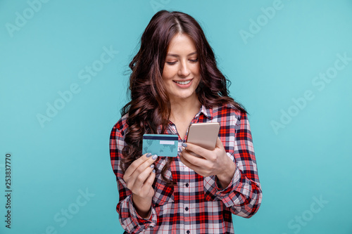 young girl isolated over blue background using mobile phone holding credit card.