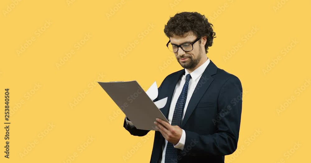 Attractive smiled Caucasian businessman in the glasses, suit and tie looking at the documents in his hands and being satisfied on the yellow back wall screen. . Attractive smiled Caucasian businessman in the glasses, suit and tie looking at the documents in his hands and being satisfied on the yellow back wall screen. .