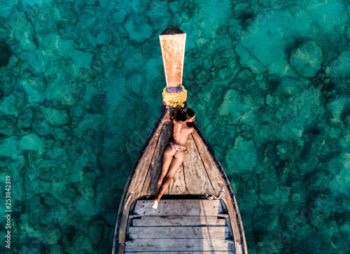Aerial drone shot of a young brunette woman without bra in the front of a wooden boat. Crystal clear water and corals at a tropical island and amazing beach.