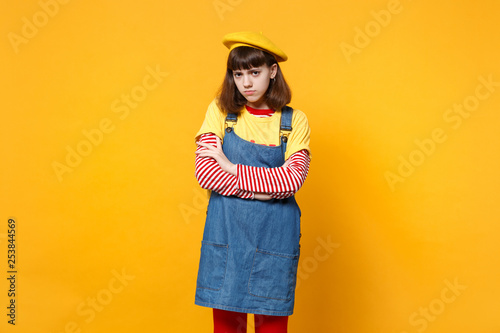 Portrait of Concerned offended girl teenager in french beret denim sundress holding hands folded isolated on yellow background in studio. People sincere emotions lifestyle concept. Mock up copy space.