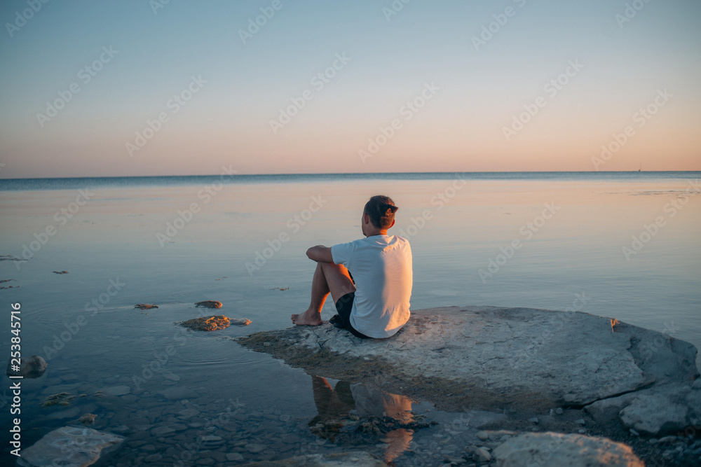 A young man sits on a stone by the sea.