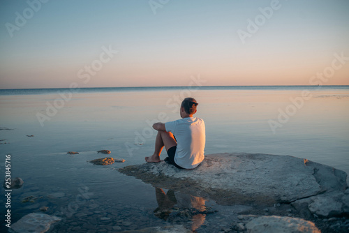 A young man sits on a stone by the sea.