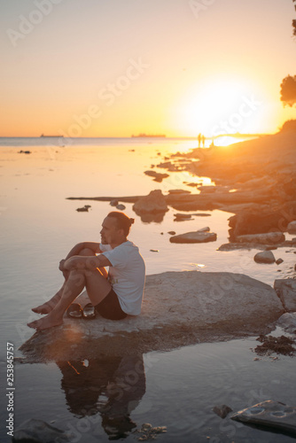 A young man sits on a stone by the sea.