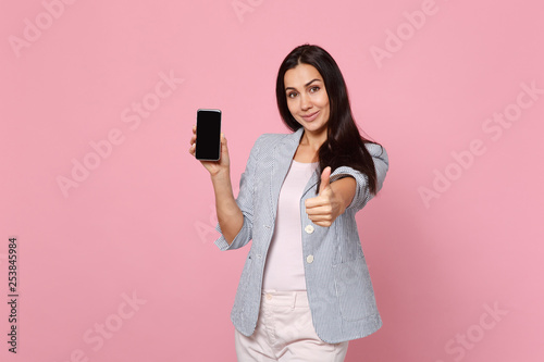 Smiling young woman in striped jacket showing thumb up, holding mobile phone with blank empty screen isolated on pink pastel background. People sincere emotions, lifestyle concept. Mock up copy space.