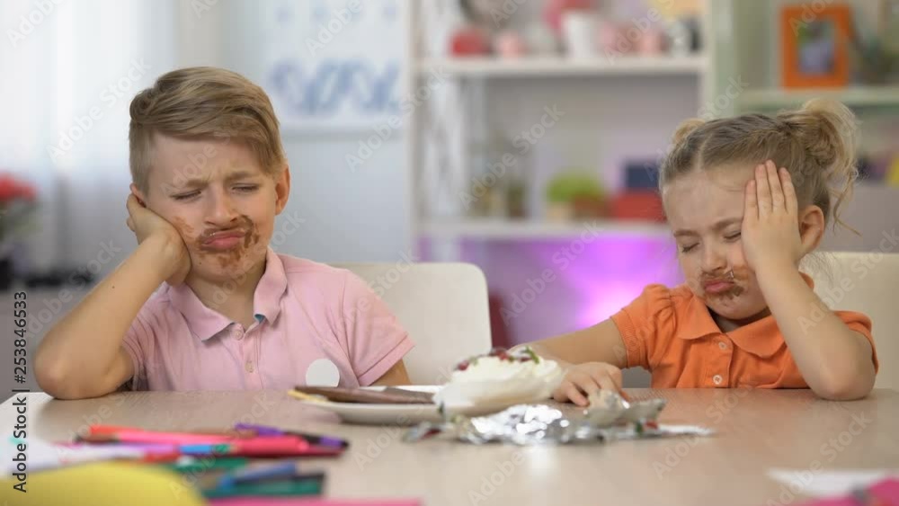 Two children smeared with chocolate sitting table, stomach pain, overeating Two children smeared with chocolate sitting table, stomach pain, overeating