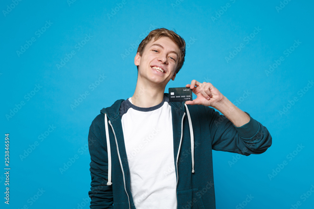 Fototapeta premium Portrait of cheerful young man in casual clothes holding credit bank card isolated on blue wall background in studio. People sincere emotions, lifestyle concept. Mock up copy space. Advertising area.