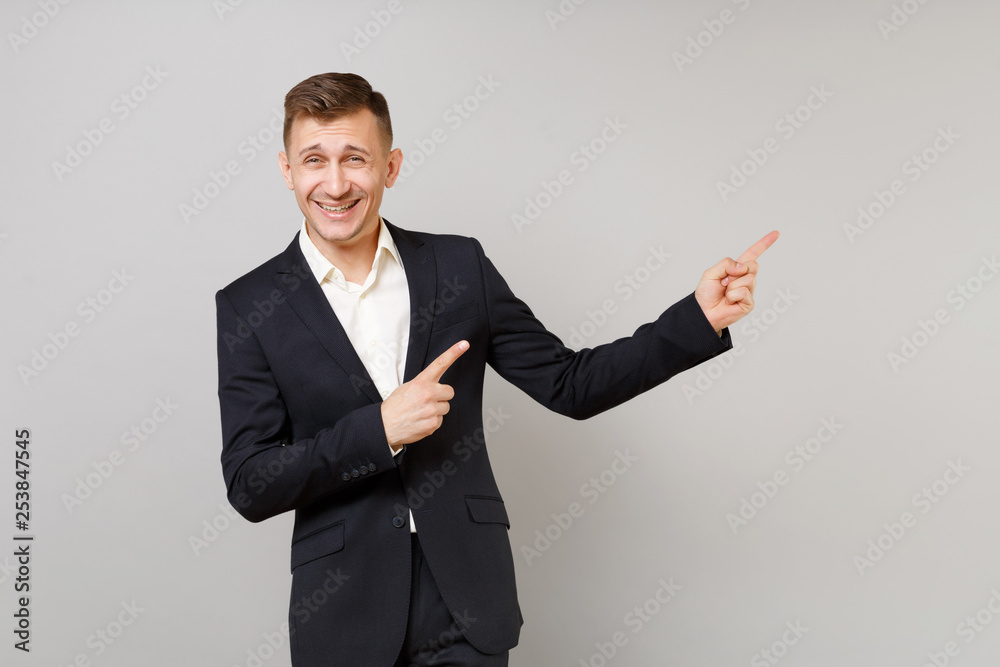Portrait of Cheerful young business man in classic black suit, shirt pointing index fingers aside isolated on grey background in studio. Achievement career wealth business concept. Mock up copy space. Portrait of Cheerful young business man in classic black suit, shirt pointing index fingers aside isolated on grey background in studio. Achievement career wealth business concept. Mock up copy space.