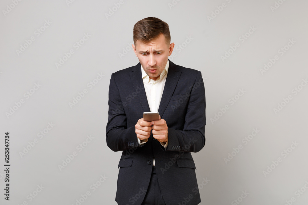Shocked concerned young business man in classic black suit using mobile phone, typing sms message isolated on grey background in studio. Achievement career wealth business concept. Mock up copy space. Shocked concerned young business man in classic black suit using mobile phone, typing sms message isolated on grey background in studio. Achievement career wealth business concept. Mock up copy space.