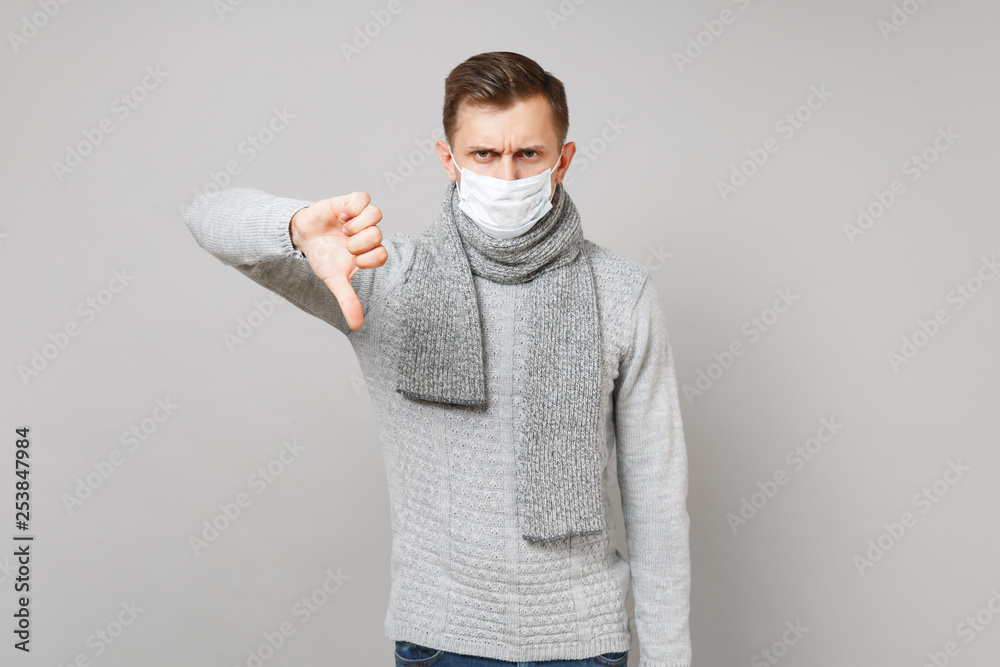 Dissatisfied young man in gray sweater, scarf sterile face mask showing thumb down isolated on grey background. Healthy lifestyle, ill sick disease treatment, cold season concept. Mock up copy space. Dissatisfied young man in gray sweater, scarf sterile face mask showing thumb down isolated on grey background. Healthy lifestyle, ill sick disease treatment, cold season concept. Mock up copy space.