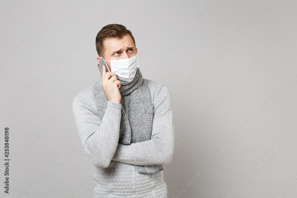 Young man in gray sweater, scarf with sterile face mask talking on mobile phone, looking aside isolated on grey background. Health, ill sick disease treatment, cold season concept. Mock up copy space. Young man in gray sweater, scarf with sterile face mask talking on mobile phone, looking aside isolated on grey background. Health, ill sick disease treatment, cold season concept. Mock up copy space.