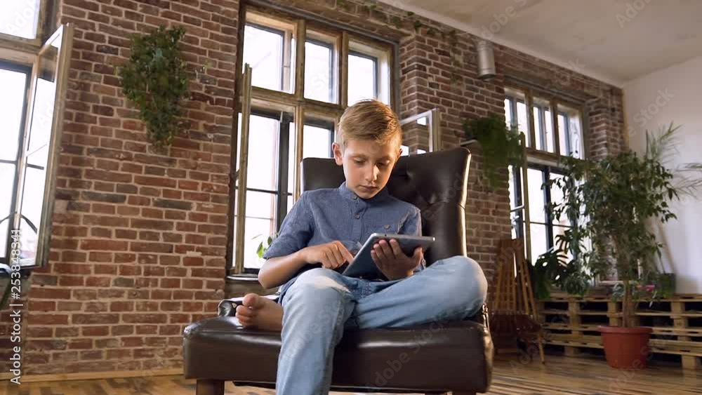 Cute caucasian boy is sitting on the armchair watching interesting movie on tablet computer . A serious teenager boy playing a games on digital tablet. Indoors. Concept of children education, science Cute caucasian boy is sitting on the armchair watching interesting movie on tablet computer . A serious teenager boy playing a games on digital tablet. Indoors. Concept of children education, science