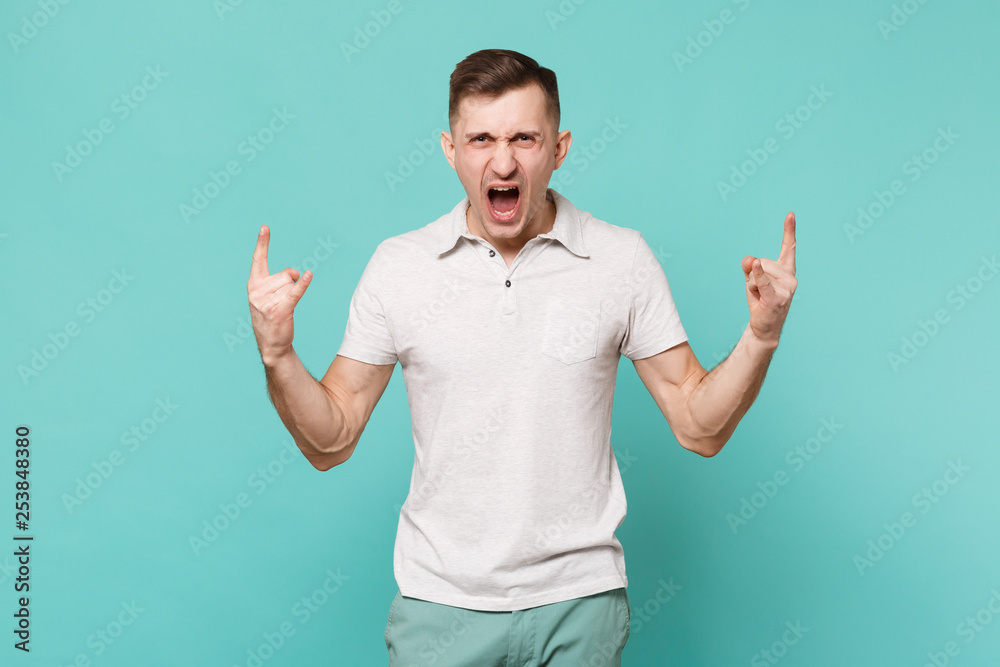 Screaming young man in casual clothes depicting horns up gesture, heavy metal rock sign isolated on blue turquoise background in studio. People sincere emotions, lifestyle concept. Mock up copy space. Screaming young man in casual clothes depicting horns up gesture, heavy metal rock sign isolated on blue turquoise background in studio. People sincere emotions, lifestyle concept. Mock up copy space.