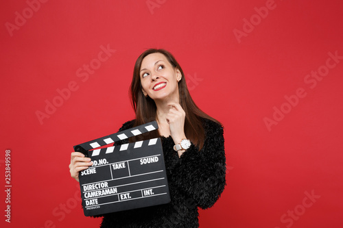 Dreamful young woman in black fur sweater looking up and holding classic black film making clapperboard isolated on red wall background. People sincere emotions, lifestyle concept. Mock up copy space.