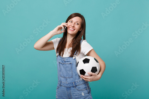Smiling pretty young woman football fan with soccer ball talking on mobile phone, conducting pleasant conversation isolated on blue turquoise background. People emotions, sport family leisure concept. © ViDi Studio