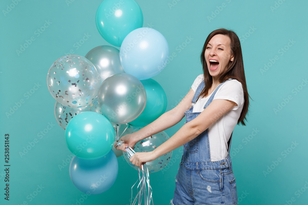 Overjoyed happy funny young woman in denim clothes screaming, celebrating and holding colorful air balloons isolated on blue turquoise wall background. Birthday holiday party, people emotions concept. Overjoyed happy funny young woman in denim clothes screaming, celebrating and holding colorful air balloons isolated on blue turquoise wall background. Birthday holiday party, people emotions concept.