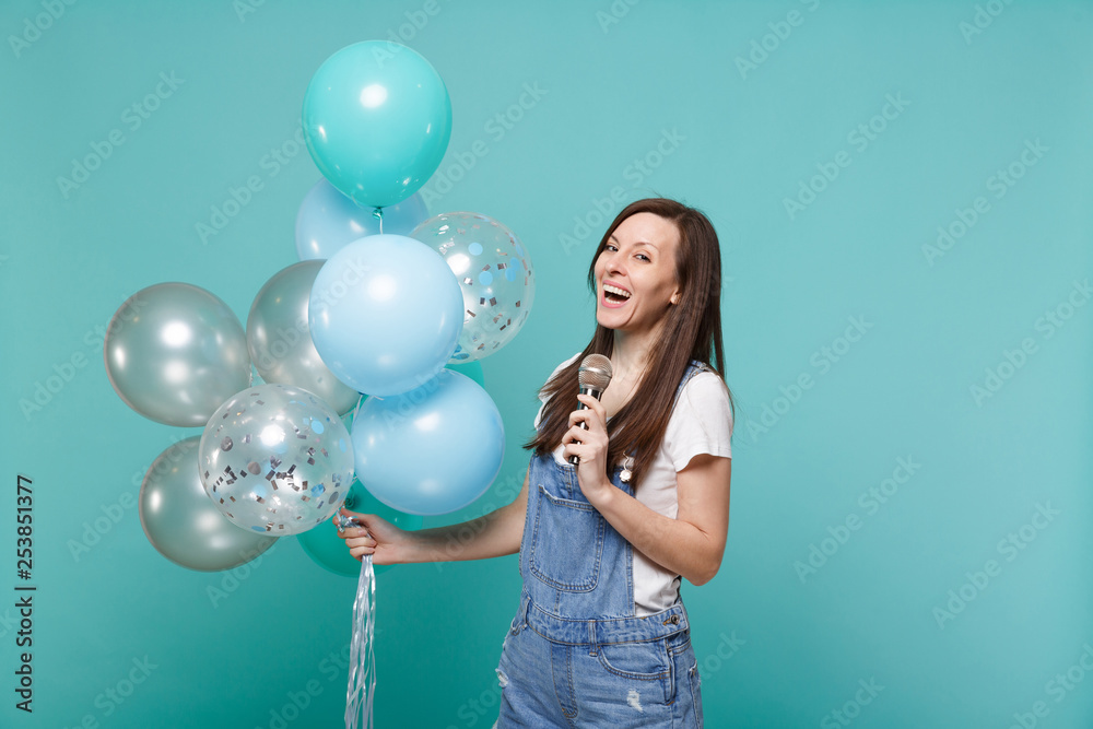 Fototapeta premium Cheerful young woman in denim clothes sing song in microphone, celebrating and holding colorful air balloons isolated on blue turquoise background. Birthday holiday party, people emotions concept.