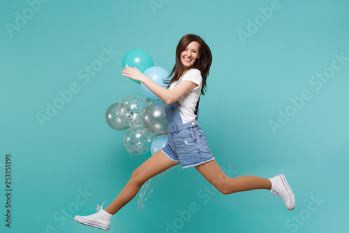 Portrait of laughing young woman in denim clothes jumping high, celebrating and holding colorful air balloons isolated on blue turquoise background. Birthday holiday party, people emotions concept.