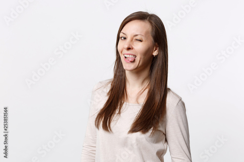 Portrait of funny cheerful blinking pretty young woman in light clothes showing tongue isolated on white wall background in studio. People sincere emotions, lifestyle concept. Mock up copy space.