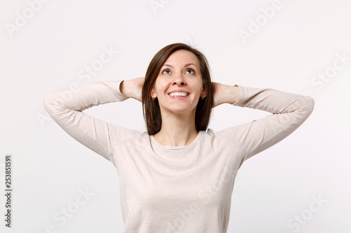 Portrait of smiling cute young woman in light clothes looking up, holding hands behind head isolated on white wall background in studio. People sincere emotions, lifestyle concept. Mock up copy space.