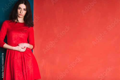 Beautiful fashion girl with dark long hair, spanish appearance and sensual face in red elegant dress posing on blue red wall in studio. Indoor lifestyle portrait of stylish woman. Fabulous brunette.
