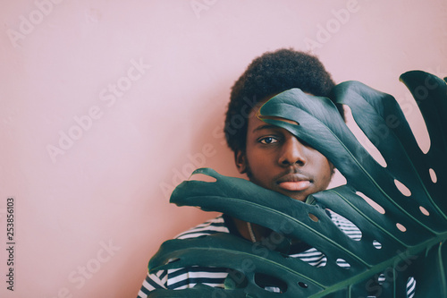 Indoor lifestyle  fashion portrait of dark-skinned african american young man standing on pink wall background with plant branch covering half face. Black nigerian male looking through palm leaves. photo