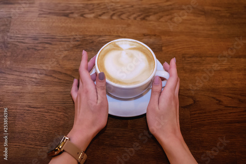 Beautiful female hands with a clock on a wooden table holding a cup of cappuccino, top view