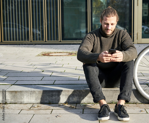 Portrait of bearded young man sitting on curb next to bicycle using cell phone