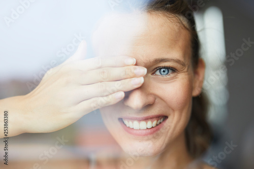 Portrait of laughing young woman behind windowpane covering her eye photo