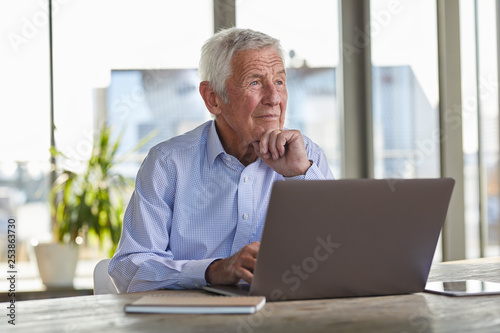 Portrait of pensive senior man sitting at table with laptop looking at distance