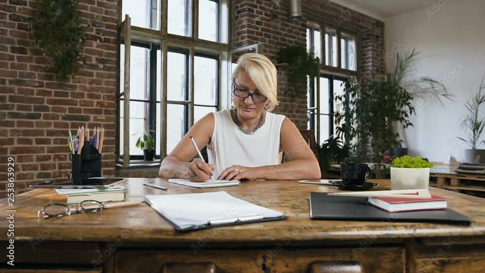 Adult attractive lady records something in the notebook while working in the home office sitting behind the desk Adult attractive lady records something in the notebook while working in the home office sitting behind the desk