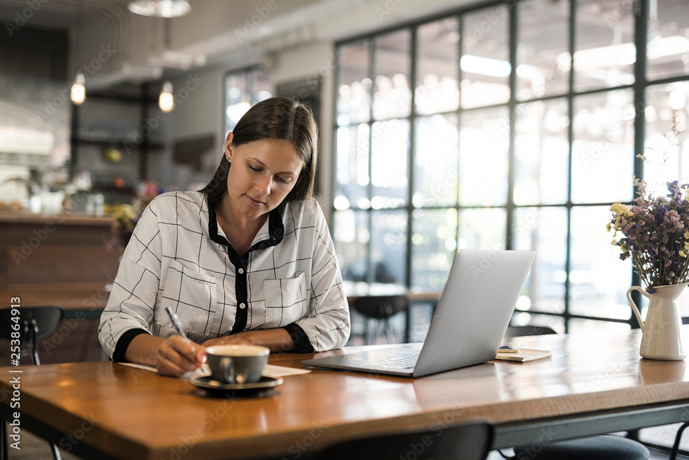 Young businesswoman in a cafe writing on paper and working with laptop on wooden table Young businesswoman in a cafe writing on paper and working with laptop on wooden table