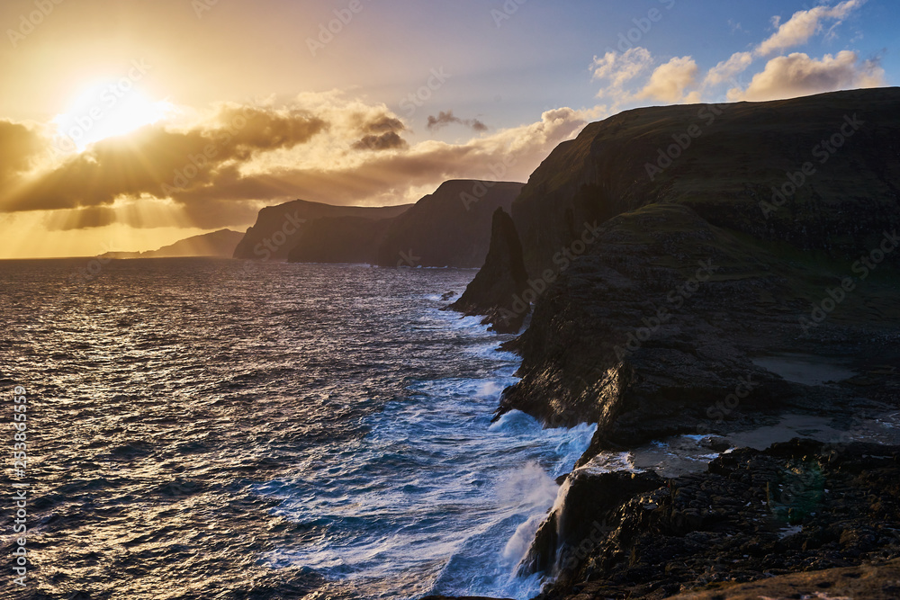 Colorful sunset over the wild rocky coastline and storm atlantic ocean in Faroe island Vagar during windy spring evening. Faroe islands, part of Kingdom of Denmark, wild scandinavian nature landscape.