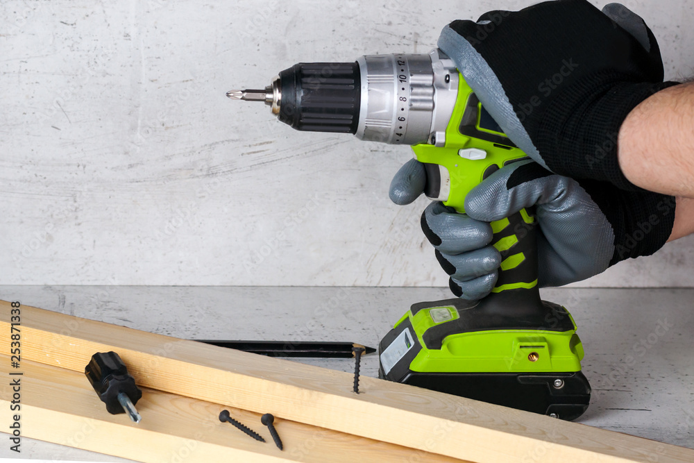The builder holds in his hand an electric screwdriver on the background of a concrete wall. Screws screw into a wooden beam. DIY