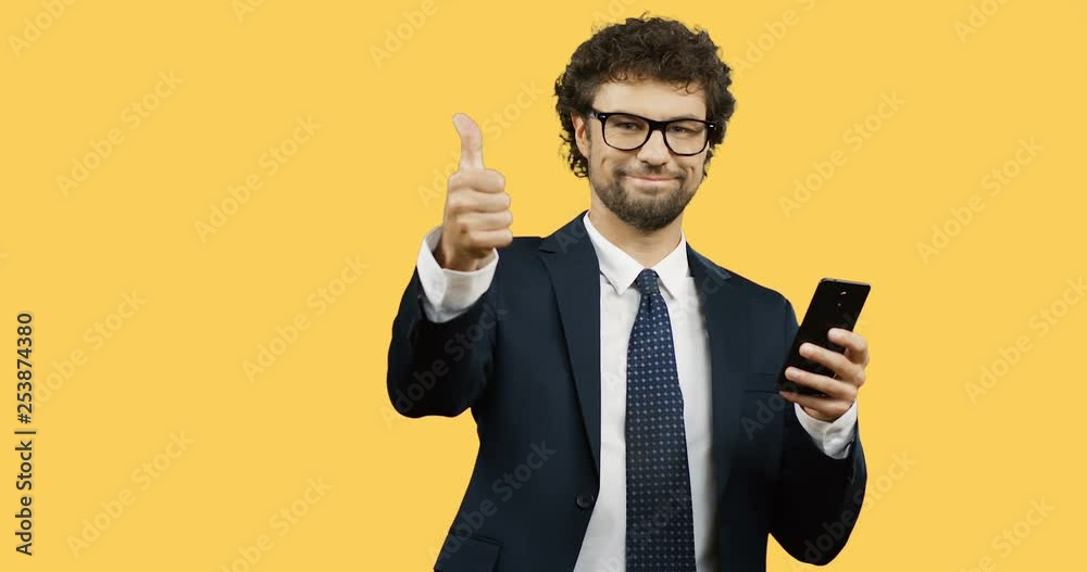 Caucasian smiled man in glasses, suit and tie giving his thumb up while scrolling and typing on the smartphone on the yellow screen background. Caucasian smiled man in glasses, suit and tie giving his thumb up while scrolling and typing on the smartphone on the yellow screen background.