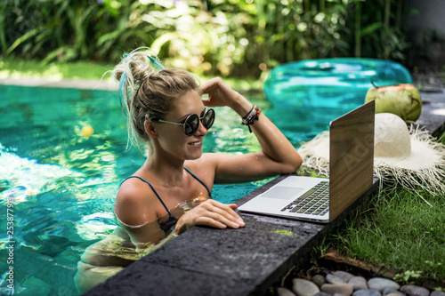 Attractive young female sitting in the swimming pool and working on a laptop. .Beautiful young female freelancer blogger working on laptop from the pool. work on vacation. photo