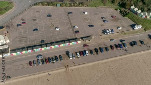 Aerial panning down over Exmouth Beach car park on a sunny day. Beach huts with multicolour doors. Cars and people us the pavements and road beside the beach. Lots of cars parked. photo