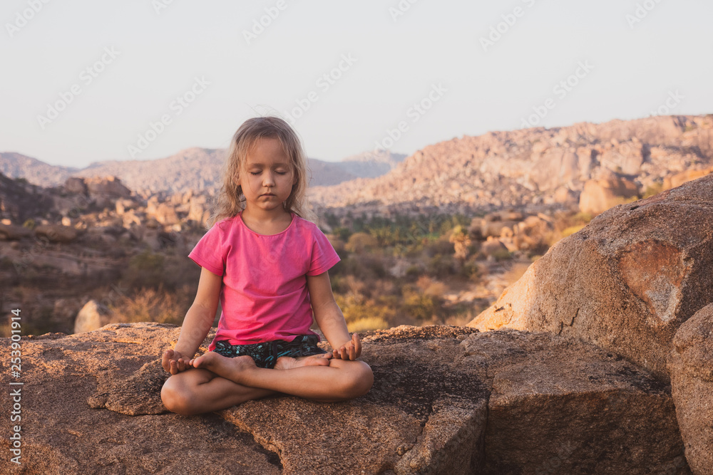 Little cute girl meditating on top of mountain. Portrait of child meditating in lotus pose on the stone