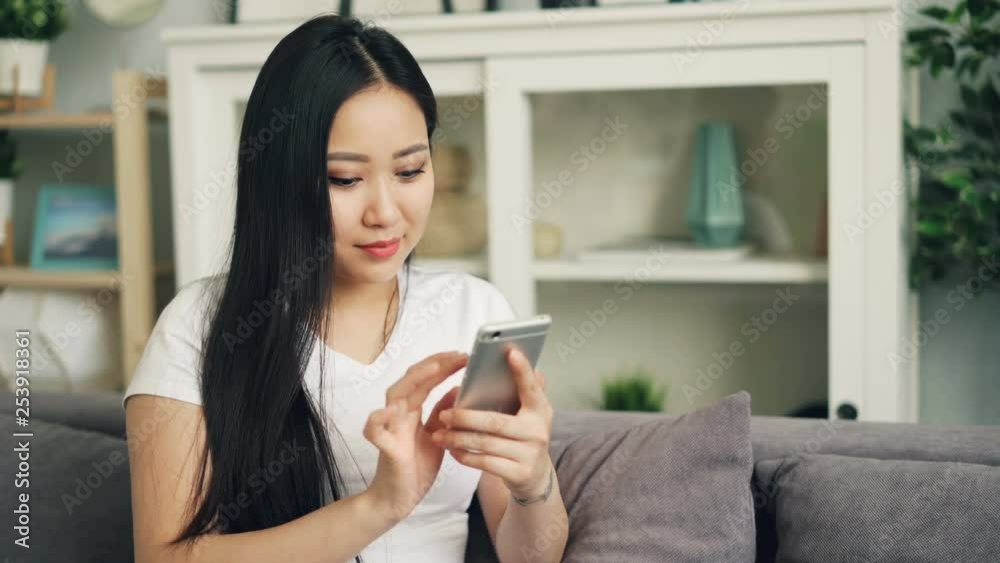 Attractive Asian woman in white T-shirt is using smartphone touching screen smiling and laughing checking social media and sending text messages. Uouth and communication concept. Attractive Asian woman in white T-shirt is using smartphone touching screen smiling and laughing checking social media and sending text messages. Uouth and communication concept.