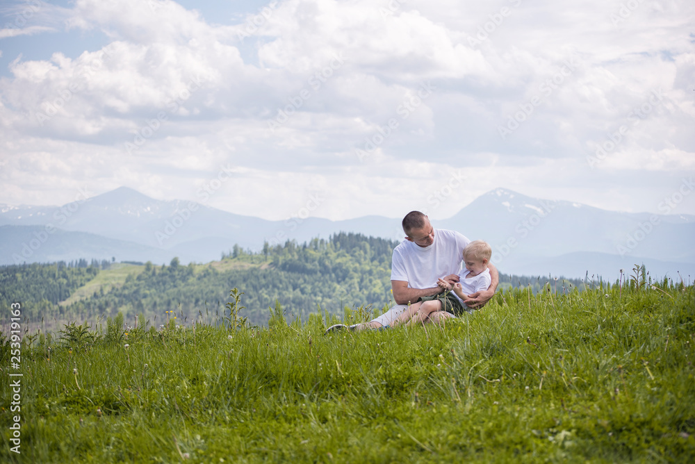 Happy father with his little son on his knees sits against a background of green forest, mountains and sky with clouds. Friendship concept