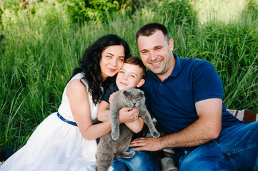 The son and parents sitting on grass hugging and holding in hands and playing with a gray cat in sunlight summer park. Happy family in the evening sun light. Mom, dad and boy at sunset.