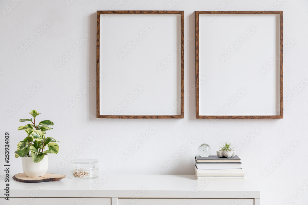 Fototapeta Minimalistic home decor of interior with two brown wooden mock up photo frames above the white shelf with books, beautiful plant in stylish pot and home accessories. White wall. Concept of mockup.